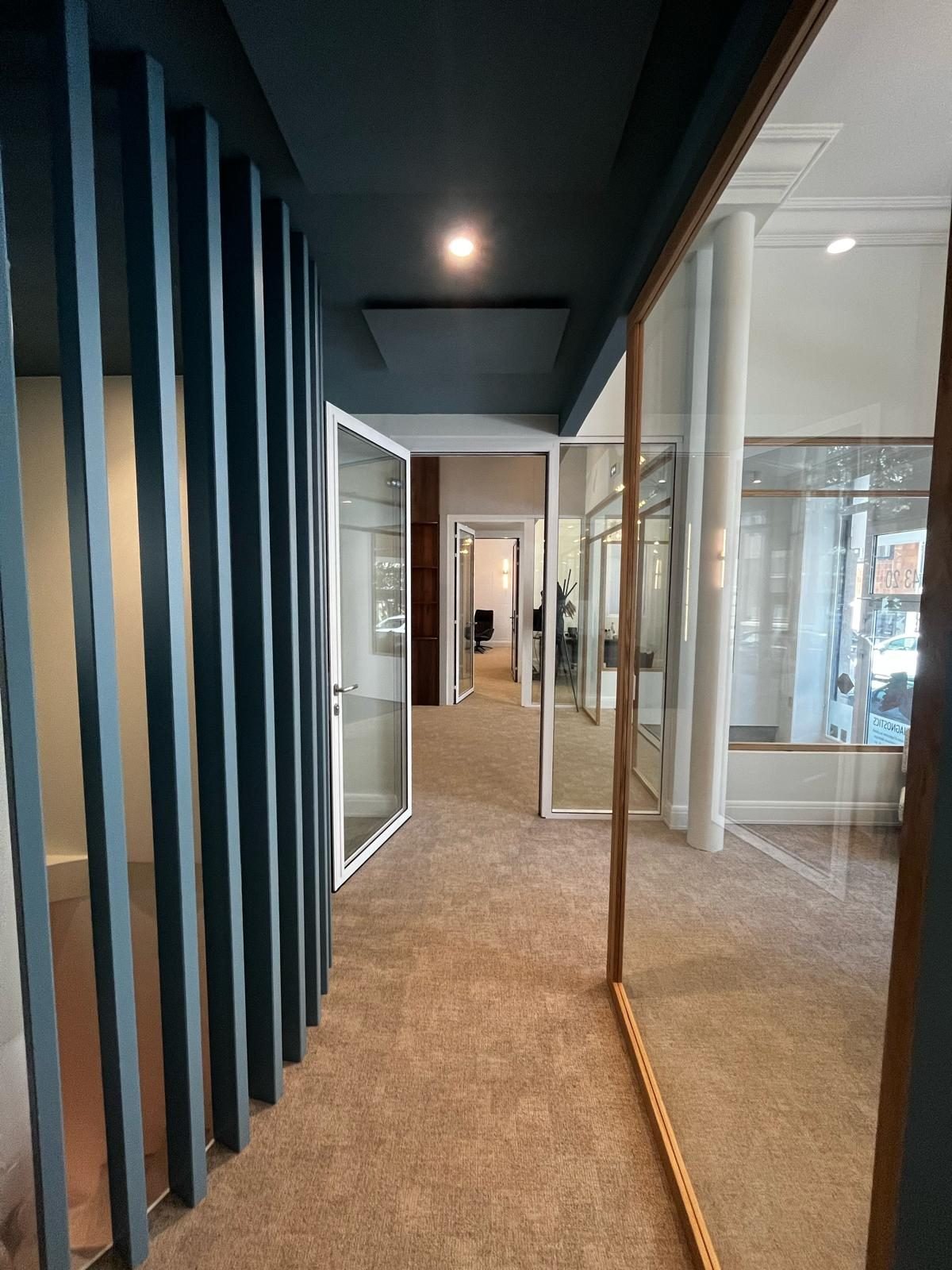 Couloir de bureaux en moquette beige entre des lames bleues et des cloisons vitrées